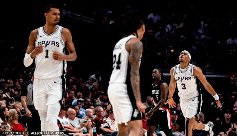 Three Spurs players in white uniforms on a basketball court during a game, with fans in the stands watching closely.